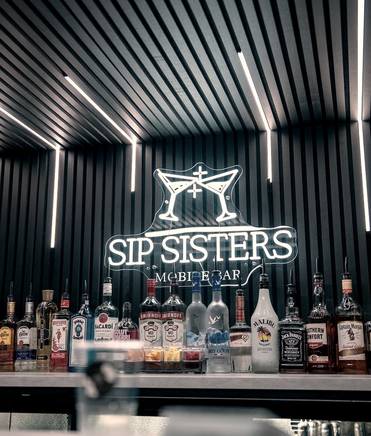Backlit Sip Sisters bar shelf with premium spirits and illuminated martini-glass sign