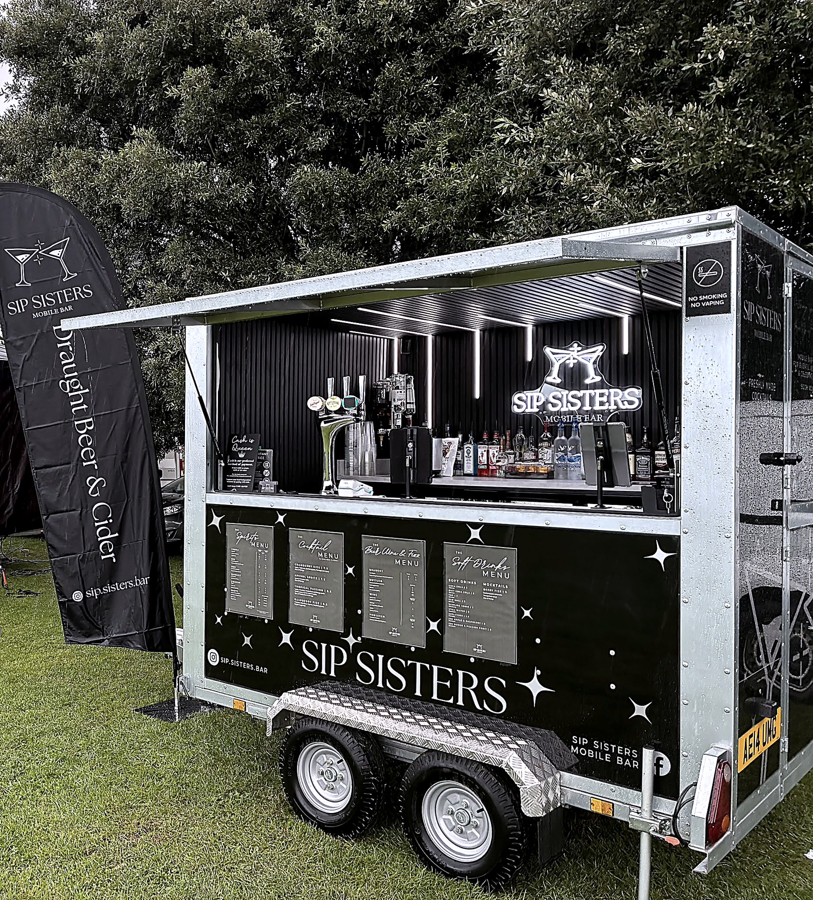 Sip Sisters bar trailer parked on grass with menu boards and open hatch